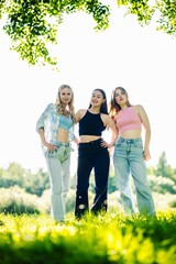 Fototapeta premium Female students smiling, standing close together in lush park setting, sharing summer friendship moment under bright sunlight