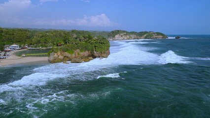 Aerial Drone view of the beach with white sand, coral rocks, hills with trees, waves from the ocean which is a tourist destination known as Drini Beach Gunung Kidul Yogyakarta