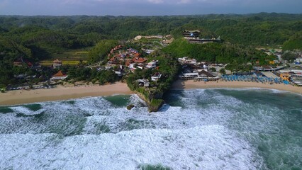 Aerial Drone view of the beach with white sand, coral rocks, hills with trees, waves from the ocean which is a tourist destination known as Drini Beach Gunung Kidul Yogyakarta