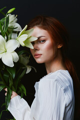 Naklejka premium Elegant portrait of a young woman with red hair holding white lilies, wearing a light blouse, against a dark background, conveying beauty and serenity