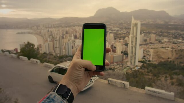 Male hand with tattoos and smart watch holds smartphone with green chroma key screen. Camper van with bike rack stand over city landscape ocean views. Travel around roadtrip lifestyle
