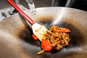 A close-up of a wok cooking stir-fried chicken and vibrant red bell peppers. The dish features a glossy sauce, highlighting the fresh ingredients and dynamic cooking process.
