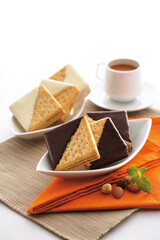 Cozy breakfast with a plate of crispy biscuits dipped in dark and white chocolate, served with a cup of steaming coffee. Garnished with nuts and mint leaves on a vibrant orange napkin.