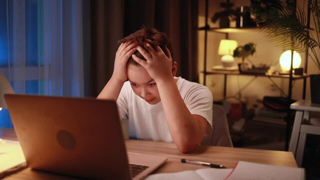 Stressed young boy holding head in frustration sitting at desk in cozy evening room emotionally reacting to laptop screen overwhelmed by online task game or remote schoolwork