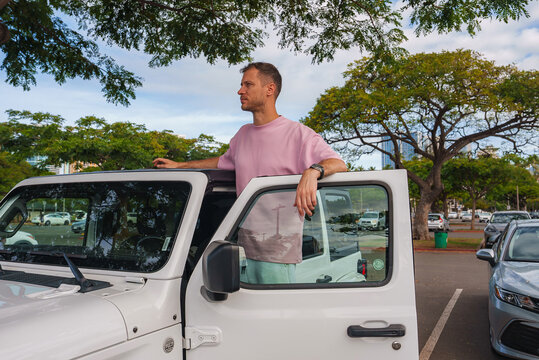 A man stands through the open door of a white Jeep in a sunny parking lot surrounded by lush green trees, with urban structures in the background. - Powered by Adobe