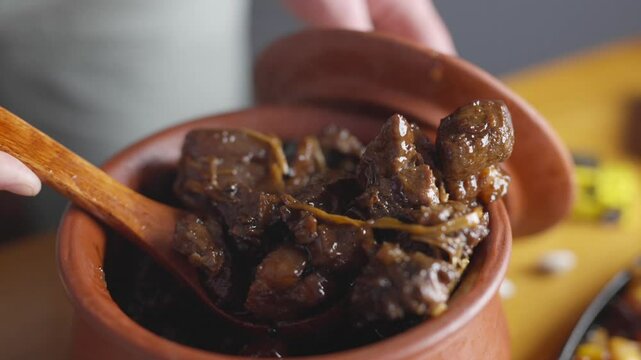 A person is holding a wooden spoon in a large pot of food. The food appears to be a stew or soup, with meat and vegetables. The pot is placed on a wooden table, and there are other items on the table