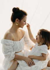 A mother and daughter share a joyful moment, dressed in white lace dresses, sitting on white...