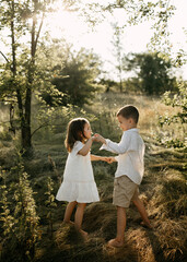 Fototapeta premium Two children holding hands and playing in a sunlit field. Brother and sister playing outdoors on summer day, barefoot.