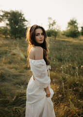 A woman in a white lace dress stands in a sunlit field, gazing softly over her shoulder.