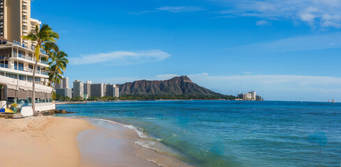 Sandy Waikiki Beach with palm trees, modern buildings, and turquoise waters, with Diamond Head crater visible under a clear blue sky.
