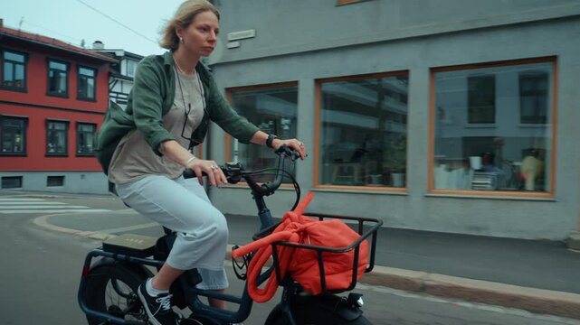 Stylish and cute young woman in casual clothes cycle through town on a cargo electric bicycle with a basket for groceries. Commuter cycling in cosy colorful old town