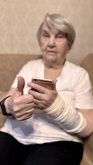 Portrait of an elderly woman of the with gray hair, sitting on the couch, looking at the camera and holding rose smartphone and sign with her hand by raising her thumb up. Close-up