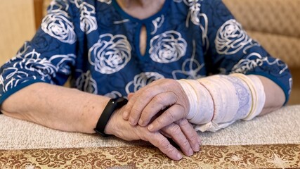 The black smart band with display touchpad and heart rate monitor. The old woman with broken arm with cast for the arm sitting in armchair touches and looks at the wristband of pulse monitor indoors
