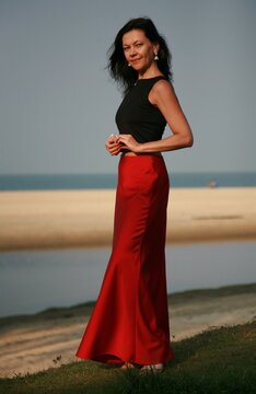 Smiling brunette woman middle aged 51 years old in red long silk skirt and indian earrings posing on beach near sea
