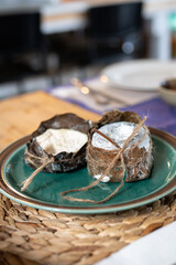 Two pieces of handmade goat cheese wrapped in fig leaves and tied with twine, presented on a ceramic plate for a rustic gourmet meal. Craft cheese production using modern technology and affinage 