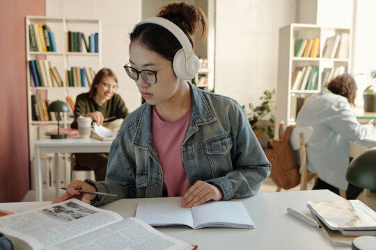 Portrait of Asian young woman wearing headphones while reading book at desk in school or college library focusing on studying for exams copy space