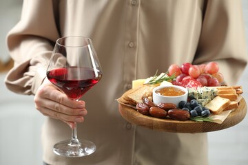 Woman holding board with different types of delicious cheese, other snacks and wine indoors, closeup