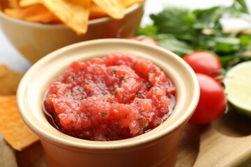 Spicy salsa sauce in bowl, nachos and ingredients on table, closeup