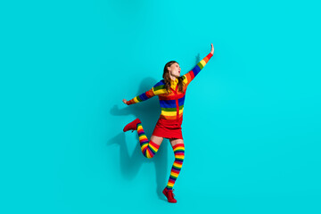 Young woman wearing vibrant rainbow clothing jumps joyfully against turquoise background reflecting...