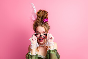 Cheerful young woman with a retro hairstyle in vintage costume posing playfully against a pink...