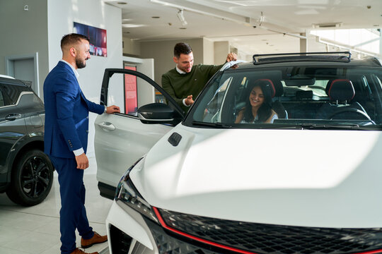 Car dealership scene with a salesman in a blue suit assisting a couple in selecting a new vehicle. The couple is exploring the interior of a white car, discussing features