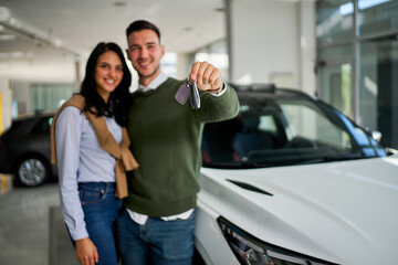 Smiling couple holding car keys, standing beside their new vehicle in a modern dealership. The scene conveys excitement and achievement in acquiring a new car