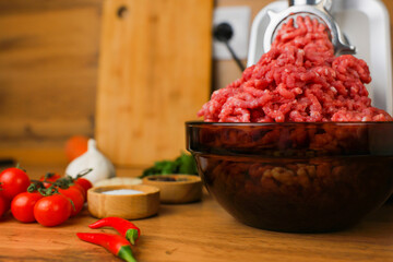 Side view on a wooden background: dark dishes with red minced meat inside, which comes out of a metal meat grinder, spices, herbs and vegetables are nearby. The concept of healthy eating, cooking