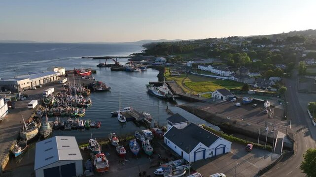 Lough Foyle Ferry, County Donegal, Ireland, June 2023. Drone pulls backwards at an angle above Greencastle harbour filled with fishing boats and a transport vessels docked in the early evening light.