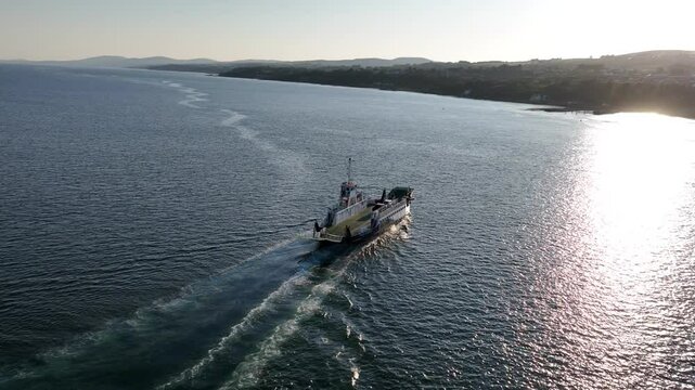 Lough Foyle Ferry, County Donegal, Ireland, June 2023. Drone quickly pulls backwards and orbits clockwise as transport vessel approaches Greencastle harbour with sun rays glistening across the water.