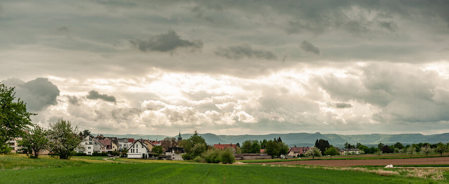 Panorama Neubausiedlung und Bauernhof vor H&uuml;gelkette der schw&auml;bischen Alb und Wolkenhimmel mit durchbrechender Sonne und Feldern und Acker bei Filderstadt-Harthausen auf den Fildern
