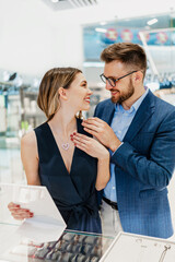 Beautiful couple enjoying in shopping at modern jewelry store. Young woman try it out gorgeous and expensive necklace and earrings. Fashion style and consumerism concept.