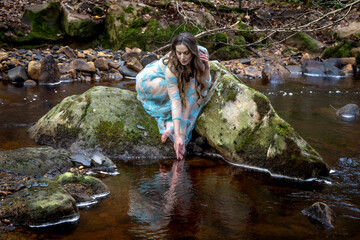 Beautiful woman in a sheer blue dress sitting on a rock, reaching down and reflected in the still water of a river © DavidOsborne