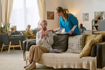 Wide shot of young nurse standing behind sofa and checking senior resident of nursing home while she enjoying crocheting