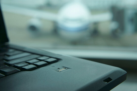 A computer laptop on a surface,in front of a window. Airport apron and aircraft seen through the window.