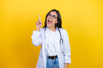 Young brunette doctor woman wearing stethoscope standing over isolated yellow background pointing finger up with successful idea. Exited and happy. Number one.