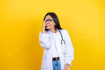 Young brunette doctor woman wearing stethoscope standing over isolated yellow background peeking in shock covering face and eyes with hand, looking through fingers with embarrassed expression