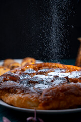 Banana Tarte Tatin is sprinkled with powdered sugar on a white plate on a dark background. The powder is poured onto the cake. Vertical Photo
