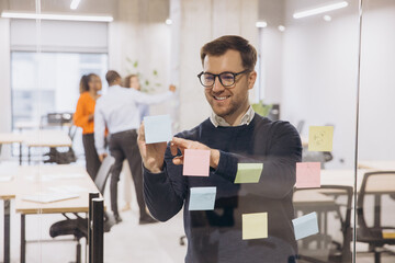 Fototapeta premium Smiling businessman using sticky notes on glass wall in a modern office, collaborating with colleagues in the background for a team project