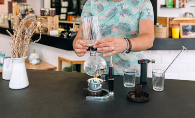 Unrecognizable barista expertly uses a Japanese siphon coffee maker in a modern cafe setting, showcasing the art of specialty coffee brewing. The process involves precision and attention to detail.