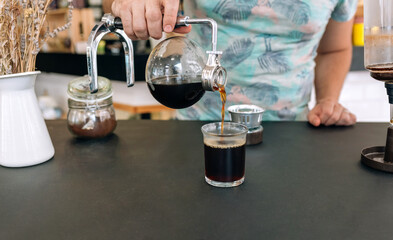 Unrecognizable barista expertly pours brewed coffee from Japanese siphon coffee maker into a glass, showcasing the art of specialty coffee preparation. The process involves much precision to details.