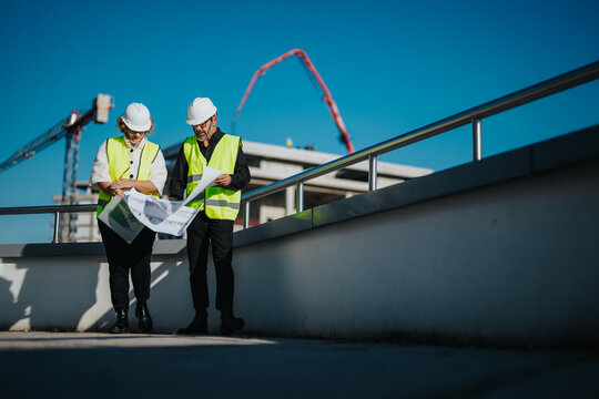 Two architects in hard hats and vests review blueprints on a construction site, symbolizing teamwork and planning. Cranes and clear sky enhance the professional atmosphere.