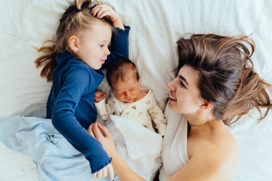 Mother lies on bed with newborn and older daughter, sharing quiet family moment. Newborn baby sleeps soundly while older sister gently watches, love, care, sibling bonding inl home setting.