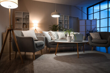Interior of cozy living room with sofa, table, armchairs and glowing lamps