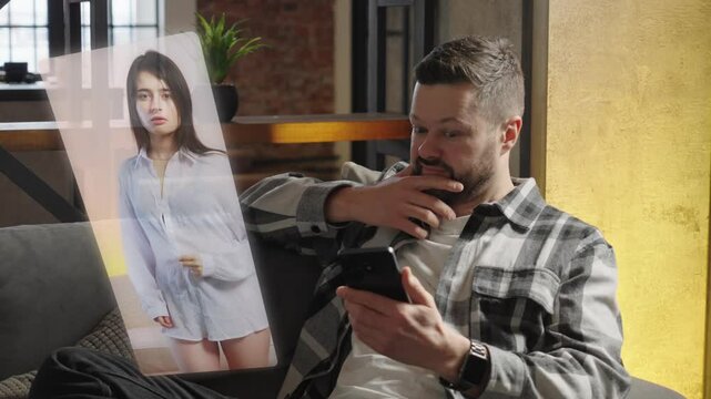 A young handsome man sits on the couch in his apartment and watches a video on his smartphone of a woman unbuttoning her shirt and dancing a hot dance