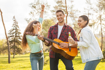 Friends gather outdoors playing guitar, singing, and enjoying a sunny afternoon with beverages.