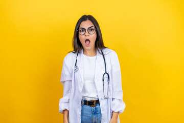 Young brunette doctor woman wearing stethoscope standing over isolated yellow background afraid and shocked with surprise and amazed expression, fear and excited face.