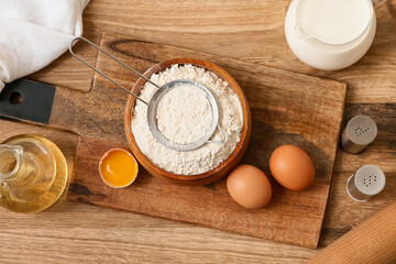 Bowl of flour with sieve, eggs, jug of oil and milk for preparing dough on wooden background