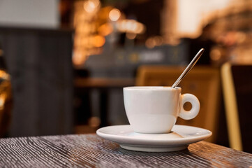 A small white cup rests on a saucer in a vibrant cafe, with a spoon gracefully placed beside it. Soft light filters through, illuminating the inviting atmosphere.