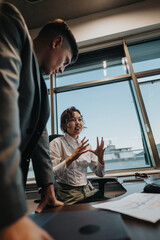 Two business professionals in a modern office setting are engaged in a discussion. Sunlight streams...