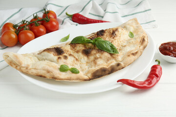Tasty calzone, basil, chili pepper, sauce and tomatoes on white wooden table, closeup